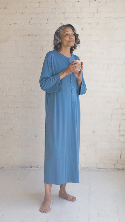 Load image into Gallery viewer, How to put on a patient gown is easy with Arkeras hospital gowns. Simple button styling in the front allow easy accessibility to chest access, perfect for breast examinations. The Henley gown is closed in the back with overlapping panels but only attched at the shoulder seam to create patient care and doctor access for all woman wearing our hospital patient gowns. Shown on our peaceful African American model in organic cotton, this beautiful ocean blue Henley patients gown is the perfect hospital gift.