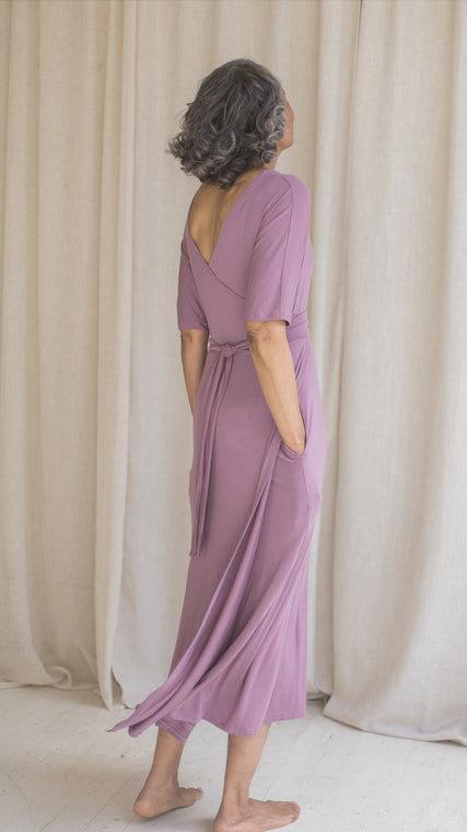 Load image into Gallery viewer, Woman wearing hospital patient gown, patient in lilac modal wrap dress, worn reversible, hand in pockets back view
